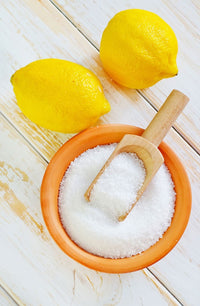 Bowl of white citric acid crystals with a wooden scoop and lemons